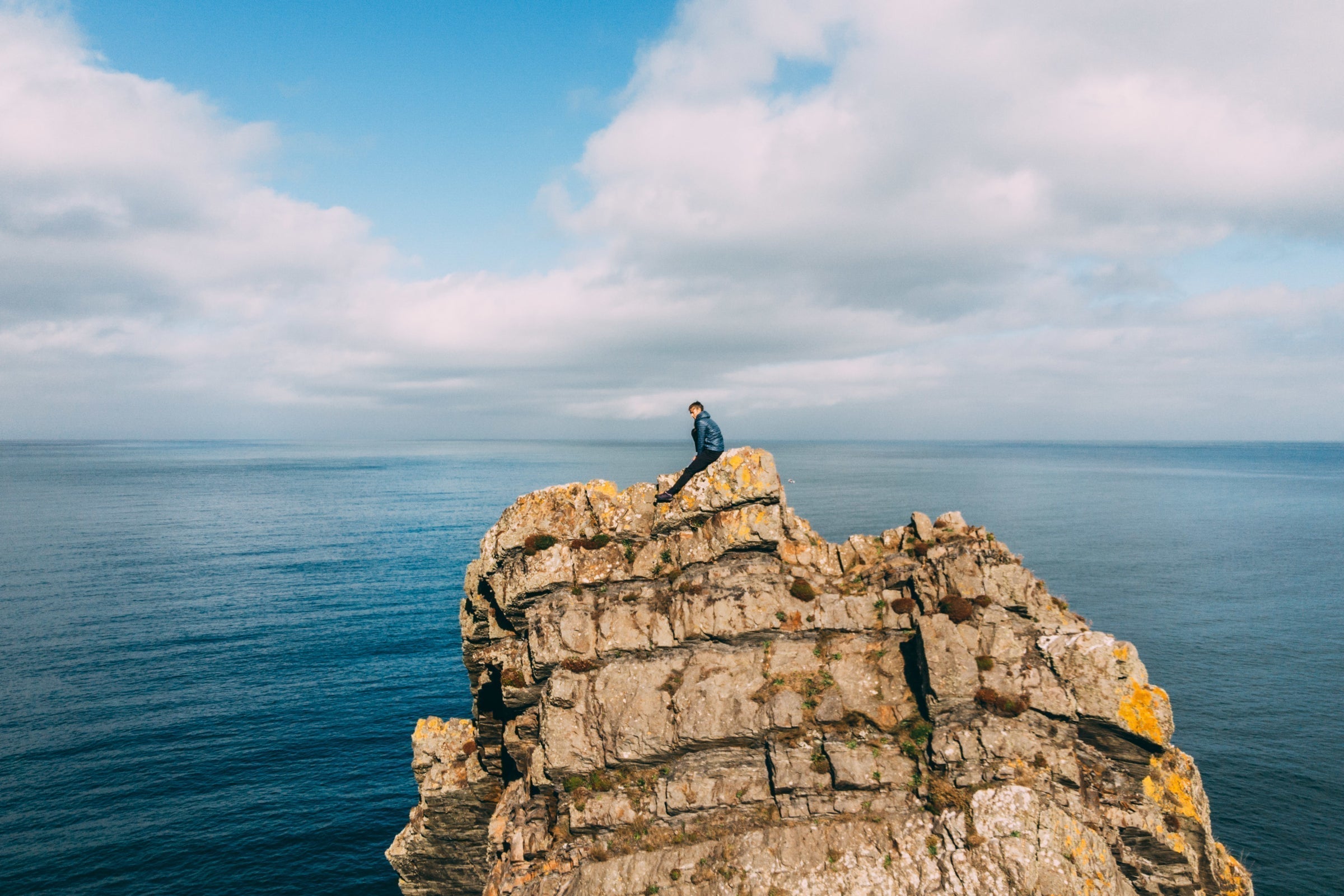 Person sitting on a rocky cliff overlooking the ocean under a partly cloudy sky