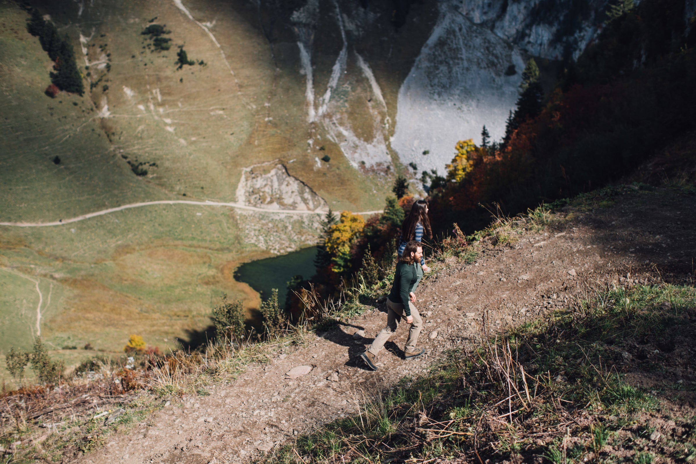 Zwei Wanderer steigen einen steilen Bergpfad hinab, mit Blick auf einen See und Wege im Tal.