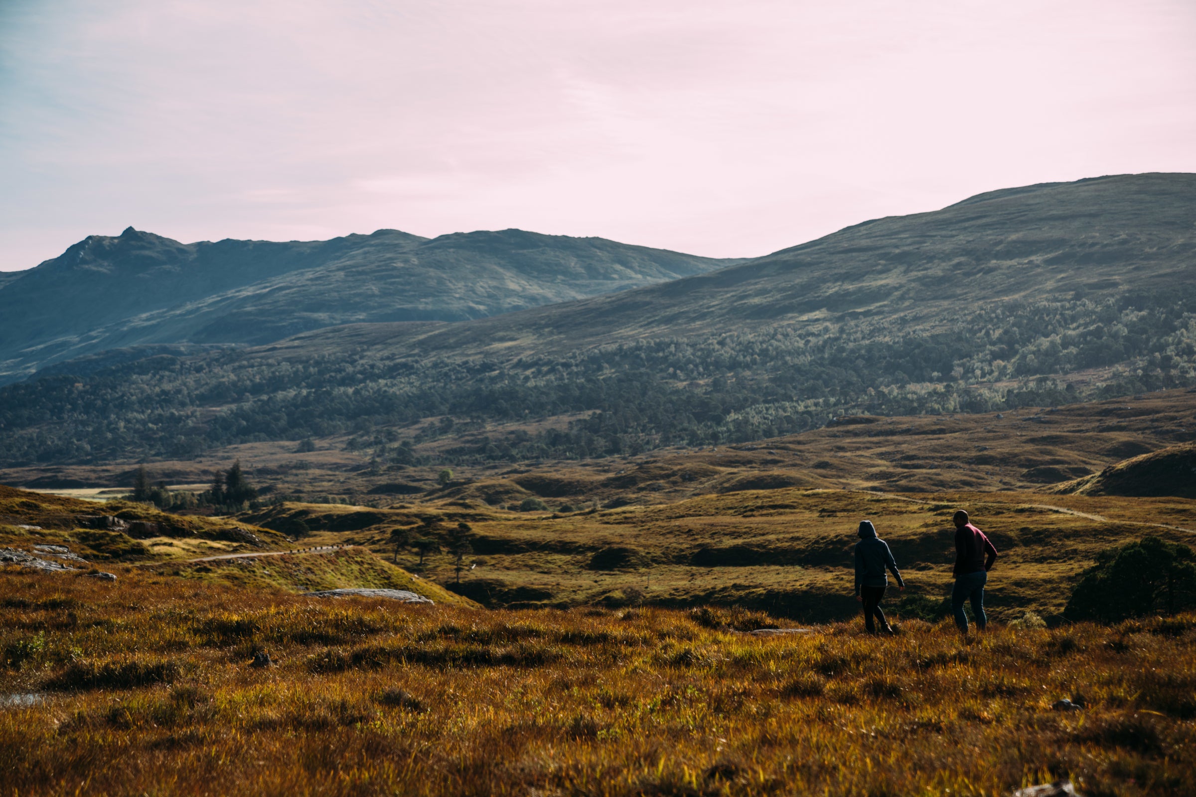 Zwei Menschen wandern über ein Moor, mit sanften Hügeln und Berggipfeln in der Ferne im warmen Sonnenlicht.