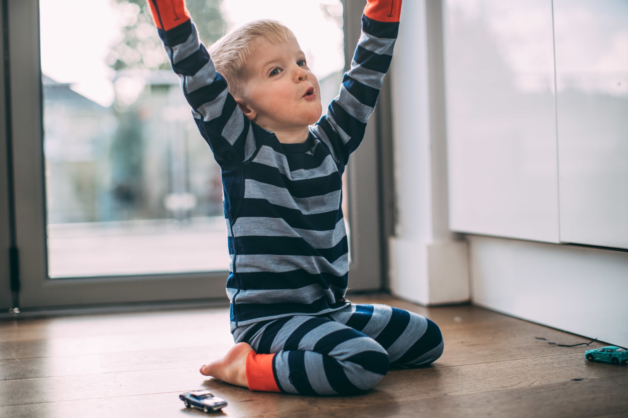 Ein kleiner Junge sitzt drinnen auf dem Boden, reißt die Arme vor Freude in die Höhe und trägt ein blaues und navy-gestreiftes Isobaa Merino Blend 200 Long Sleeve Crew und Merino Blend 200 Leggings