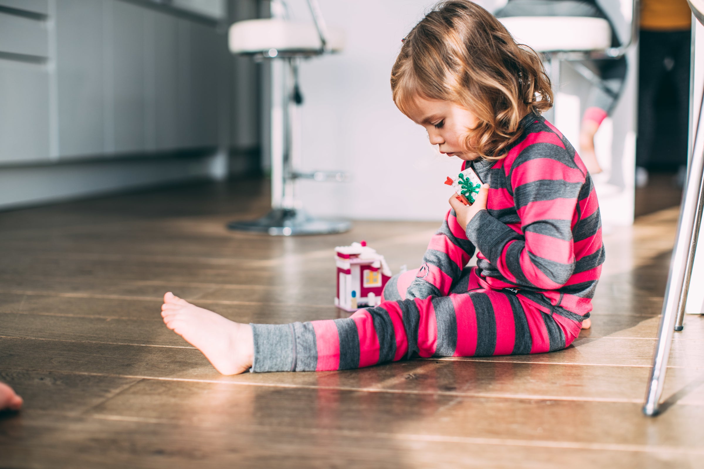 Ein kleines Mädchen spielt drinnen mit Spielzeug und trägt ein pink-grau gestreiftes Isobaa Merino Blend 200 Long Sleeve Crew und Merino Blend 200 Leggings