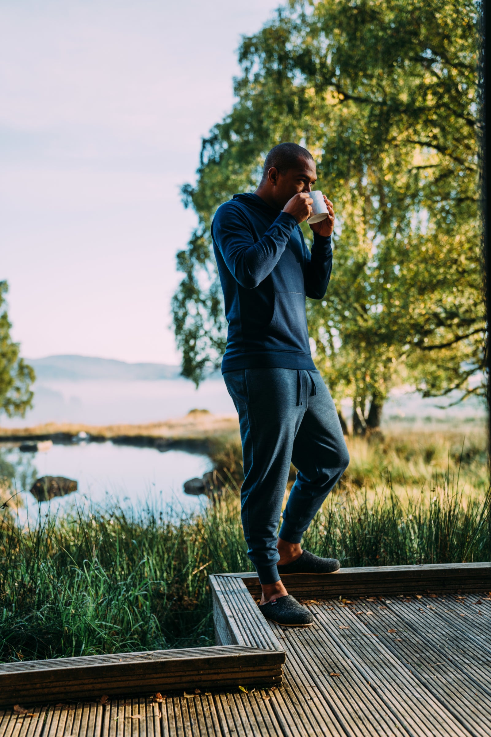 Mann genießt draußen eine Tasse Kaffee, trägt dunkelgraue Merino Blend Slippers und navyfarbenes Loungewear