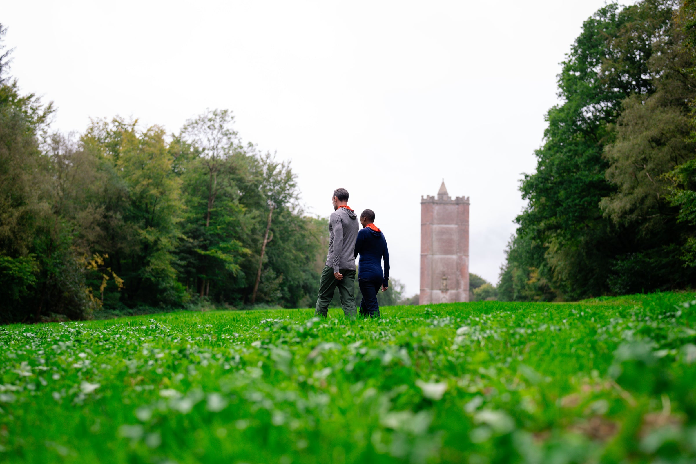 Zwei Personen gehen durch ein grünes Feld mit einem hohen Turm im Hintergrund